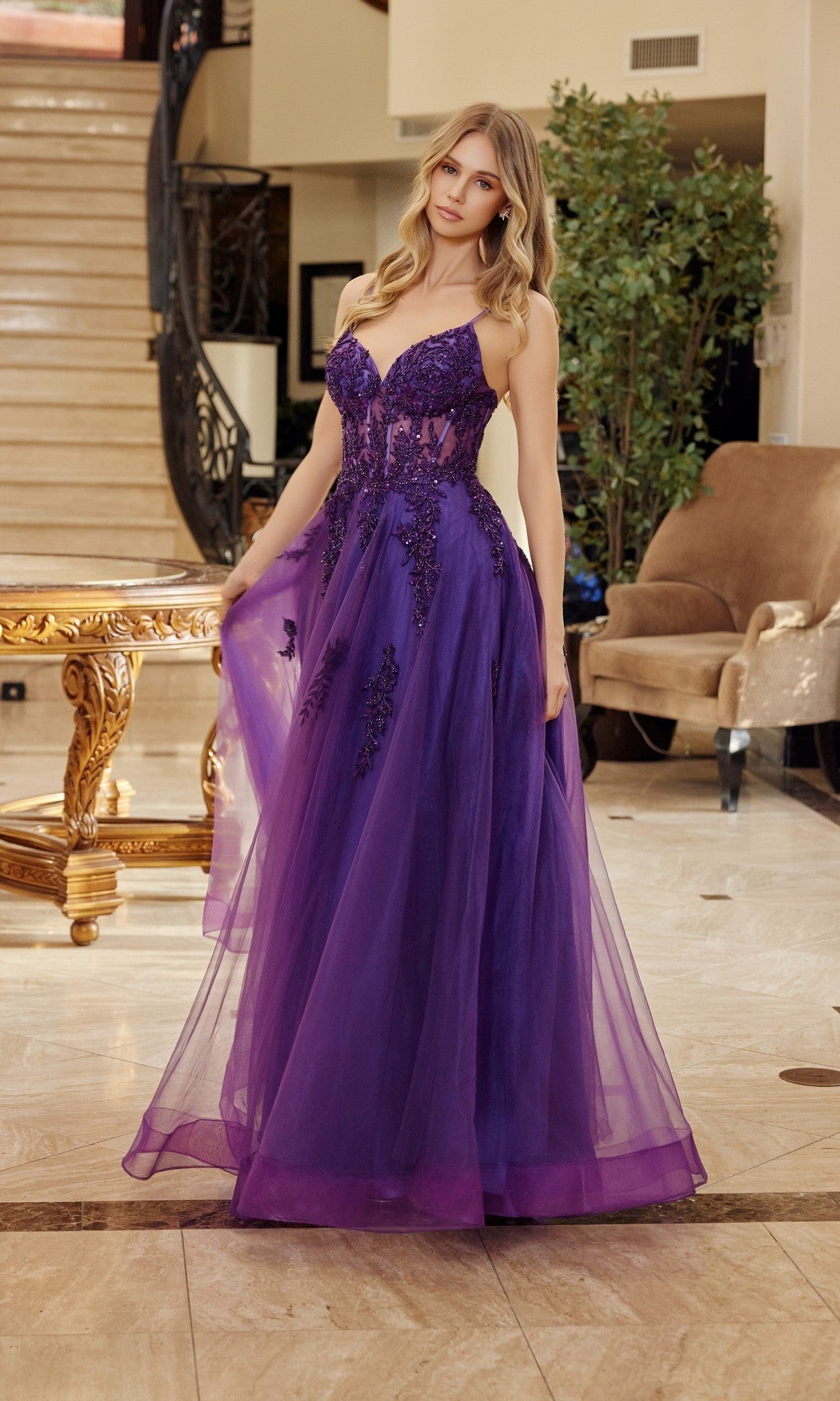 A woman stands indoors wearing the Nox Anabel Q1778 Long Prom Dress in plum, a floor-length sequin gown with floral lace accents, holding the skirt. Marble floor, staircase, and furniture appear in the background.